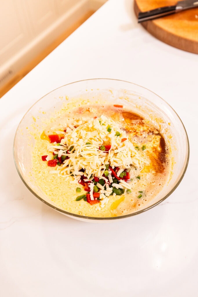 Diced peppers, cheddar cheese, and brown butter sit atop cornbread batter, ready to be mixed in.