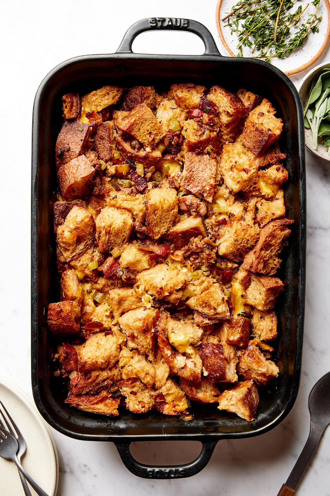 Black baking dish filled with baked sausage stuffing with herbs and plates to the side before serving.