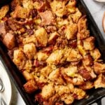 Black baking pan filled with sausage stuffing with herbs and plates to the side all on marble surface.