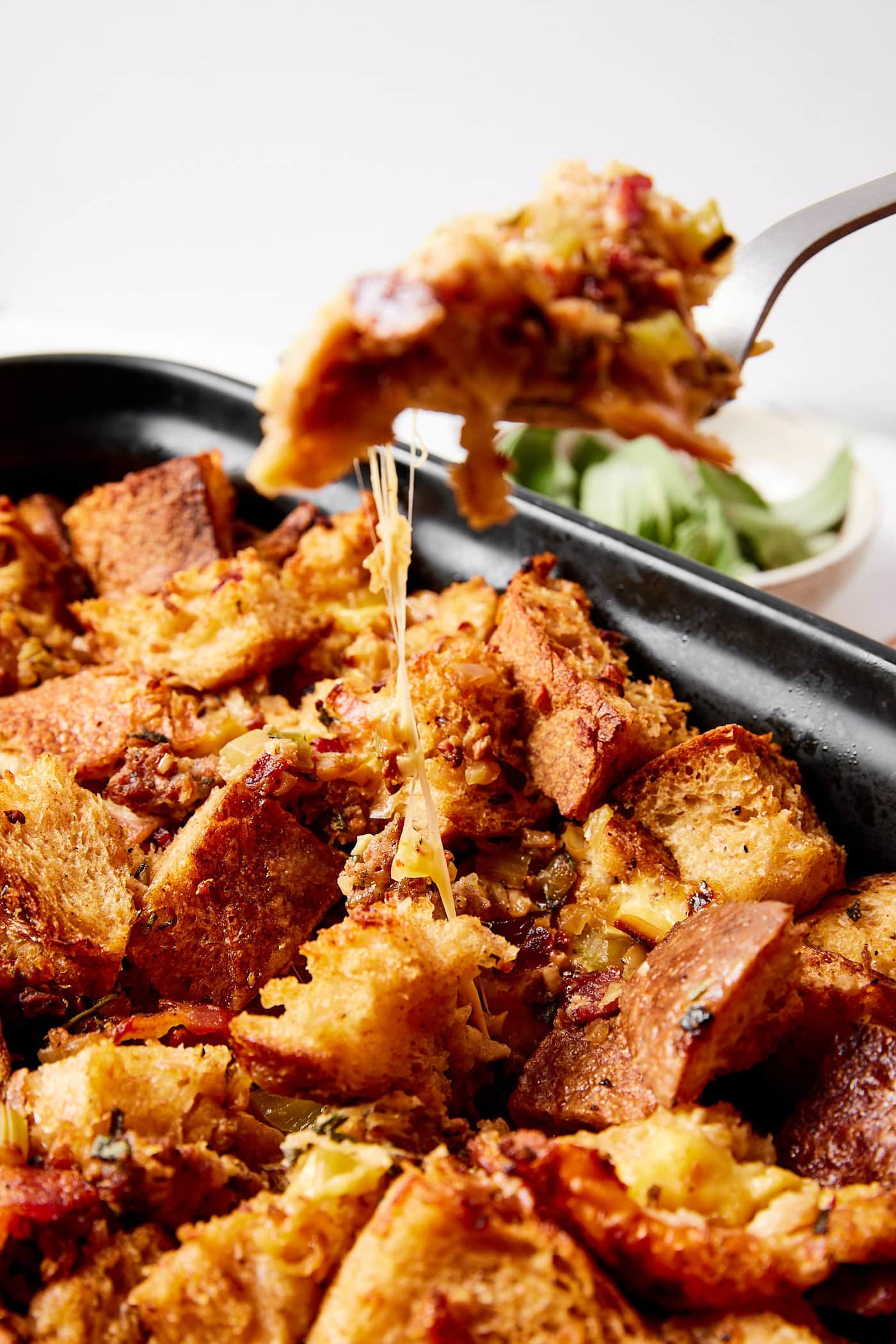 Serving of sausage stuffing on a spoon with a pull of cheese from more stuffing below in pan.