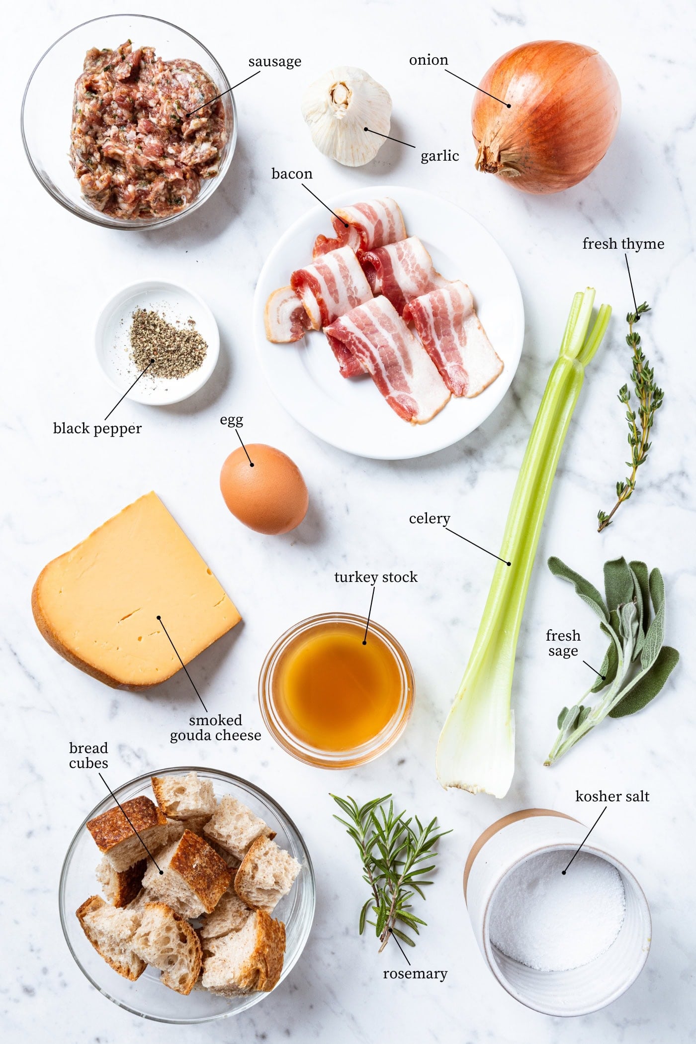 Ingredients to make bacon & cheese stuffing sit on a white marble surface. Ingredients include: sausage, garlic, onion, bacon, egg, celery, thyme, sage, black pepper, turkey stock, smoked gouda, bread, rosemary, and kosher salt.
