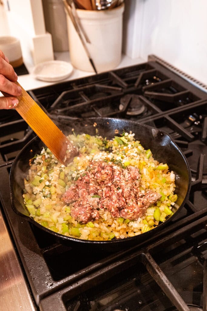 Sausage getting added to cooked onion and celery in a skillet on a stove.