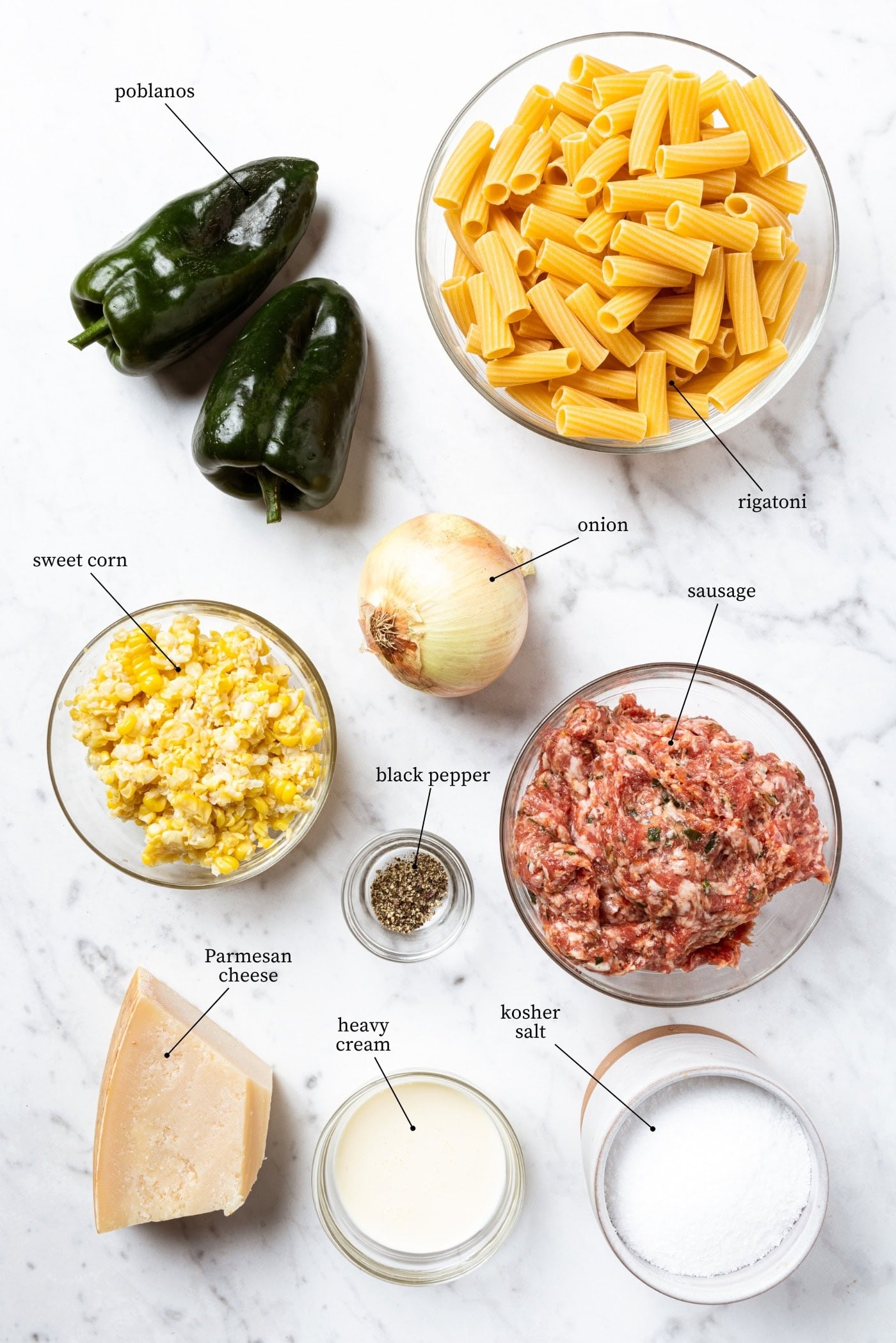 Ingredients to make poblano sweet corn pasta lie on a marble surface. Includes poblanos, pasta, onion, sweet corn, sausage, black pepper, Parmesan cheese, heavy cream, and kosher salt.