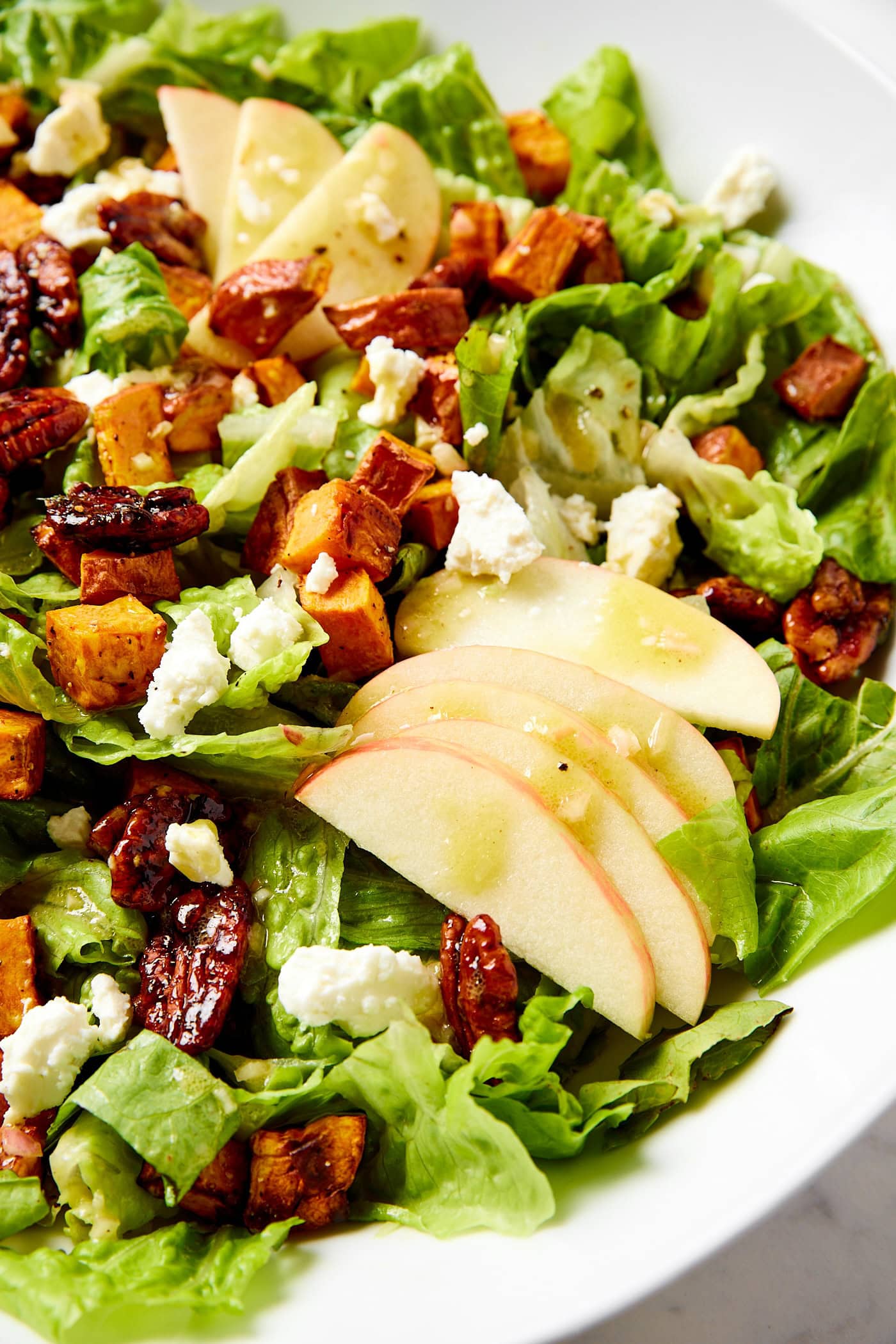 White bowl filled with sweet potato salad with apples drizzled with more dressing and candied pecans.
