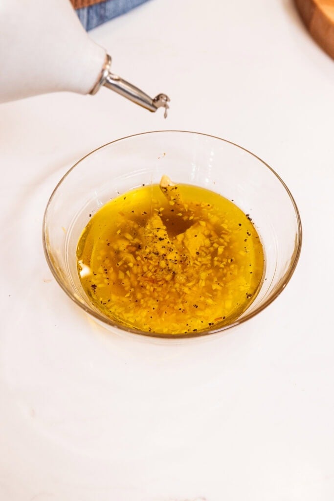 Pouring olive oil into a small bowl with other ingredients to create a vinaigrette.
