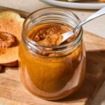 Jar filled with pumpkin butter with piece of toast in background.