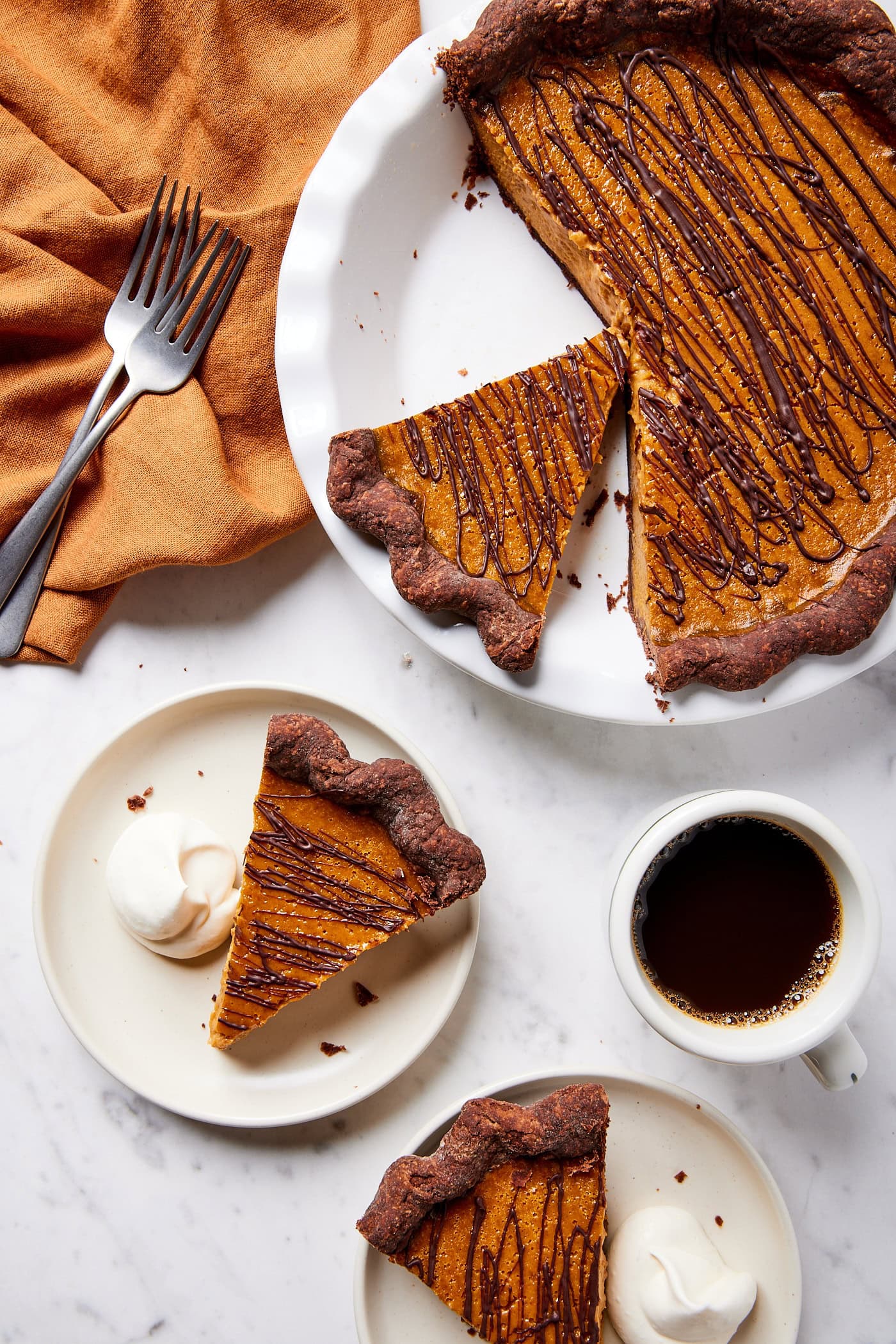 White marble surface with pumpkin pie made with a chocolate crust with slices sitting on plates with coffee and forks.