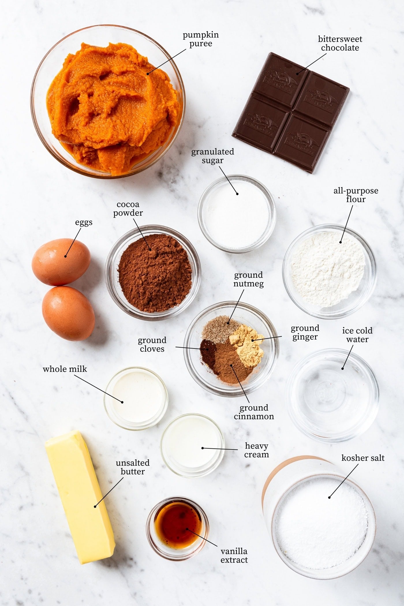 White marble surface filled with ingredients needed to make pumpkin pie with eggs, spices, flour, butter, chocolate, and more.