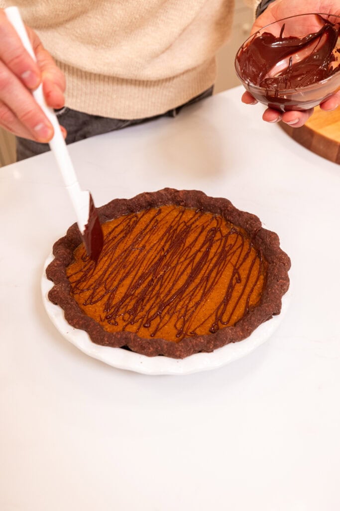 Hand drizzling chocolate on top of pumpkin pie with chocolate crust.