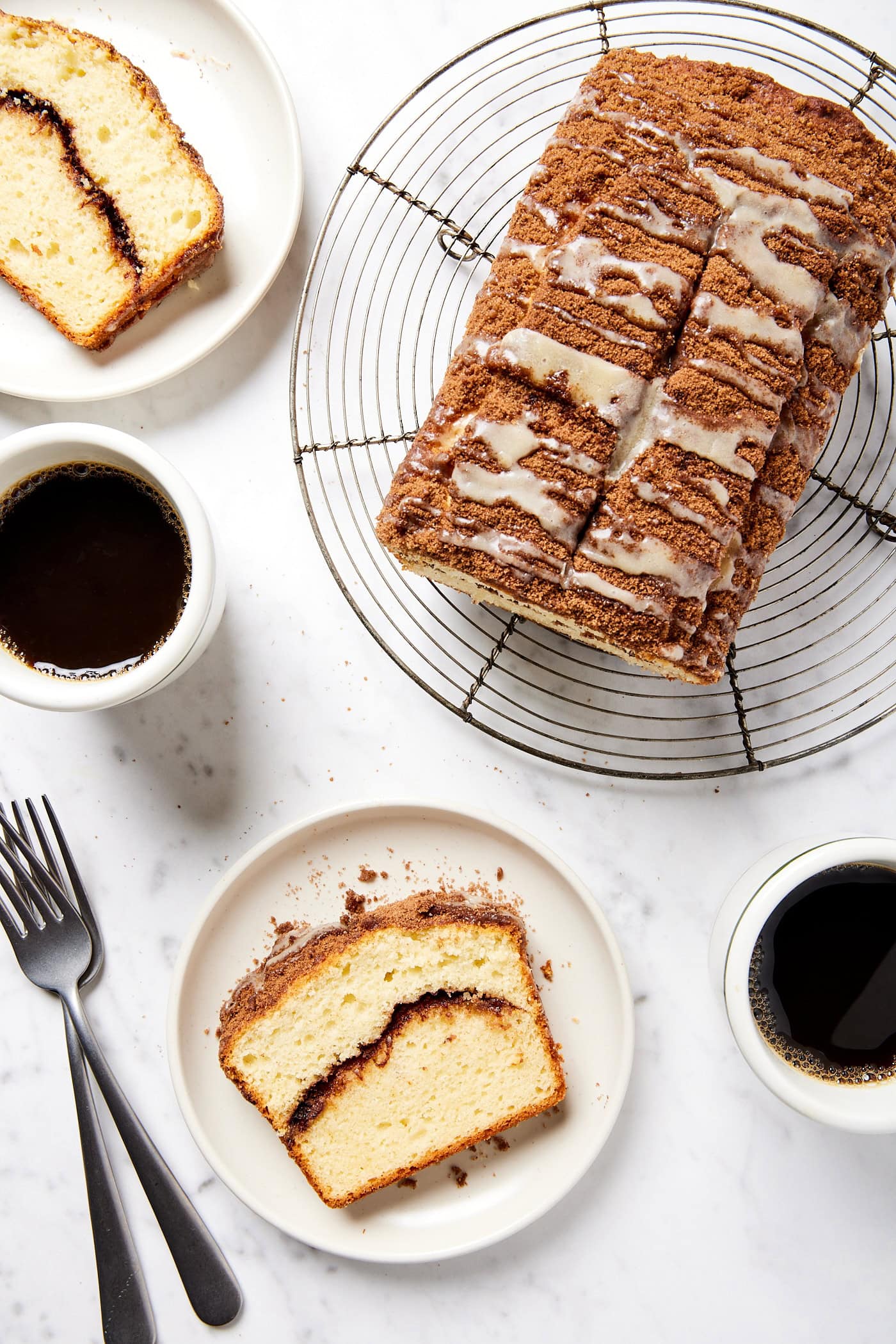 White marble surface with loaf of coffee cake with slices on plates and cups of coffee.