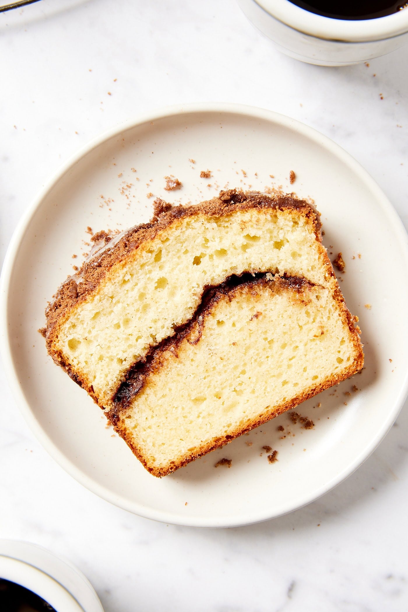 White plate with slice of coffee cake with ribbon of cinnamon sugar in the middle with cups of coffee to the side.