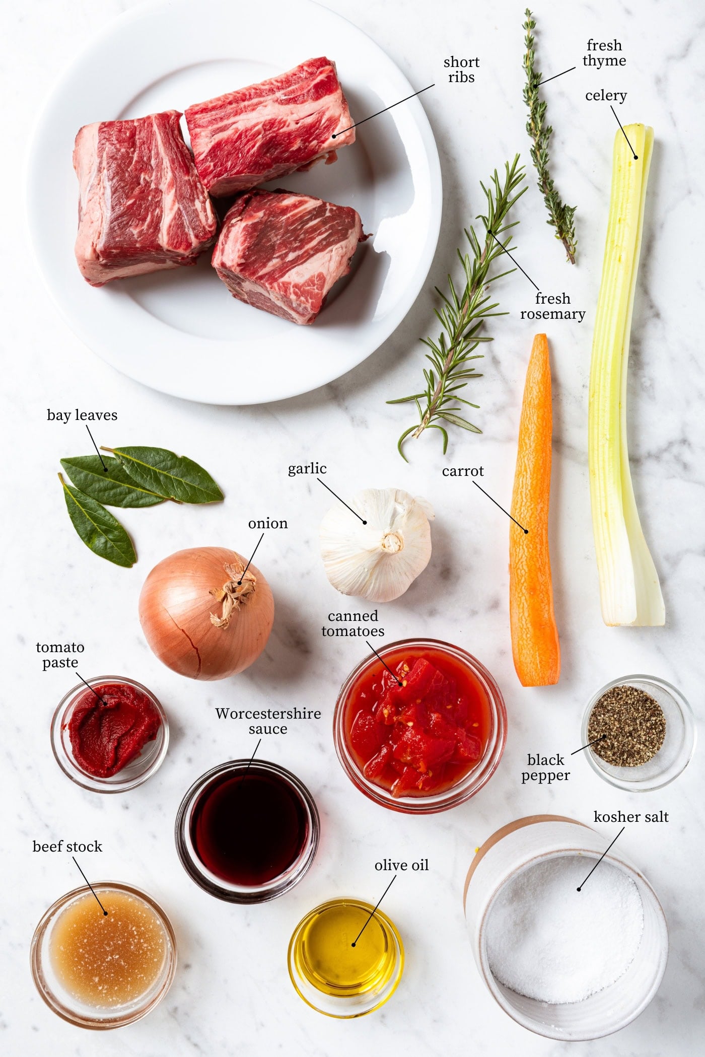 White marble surface filled with ingredients needed for short ribs including short ribs, herbs, carrots, celery, and more.