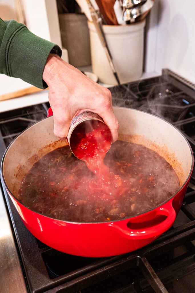 Adding canned tomatoes to a wine sauce in a large Dutch oven.
