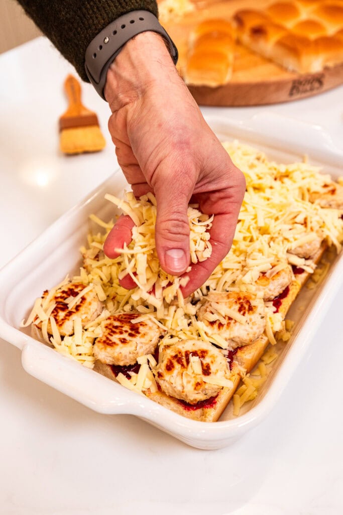 Sprinkling fresh cheddar cheese over meatballs on sweet rolls.