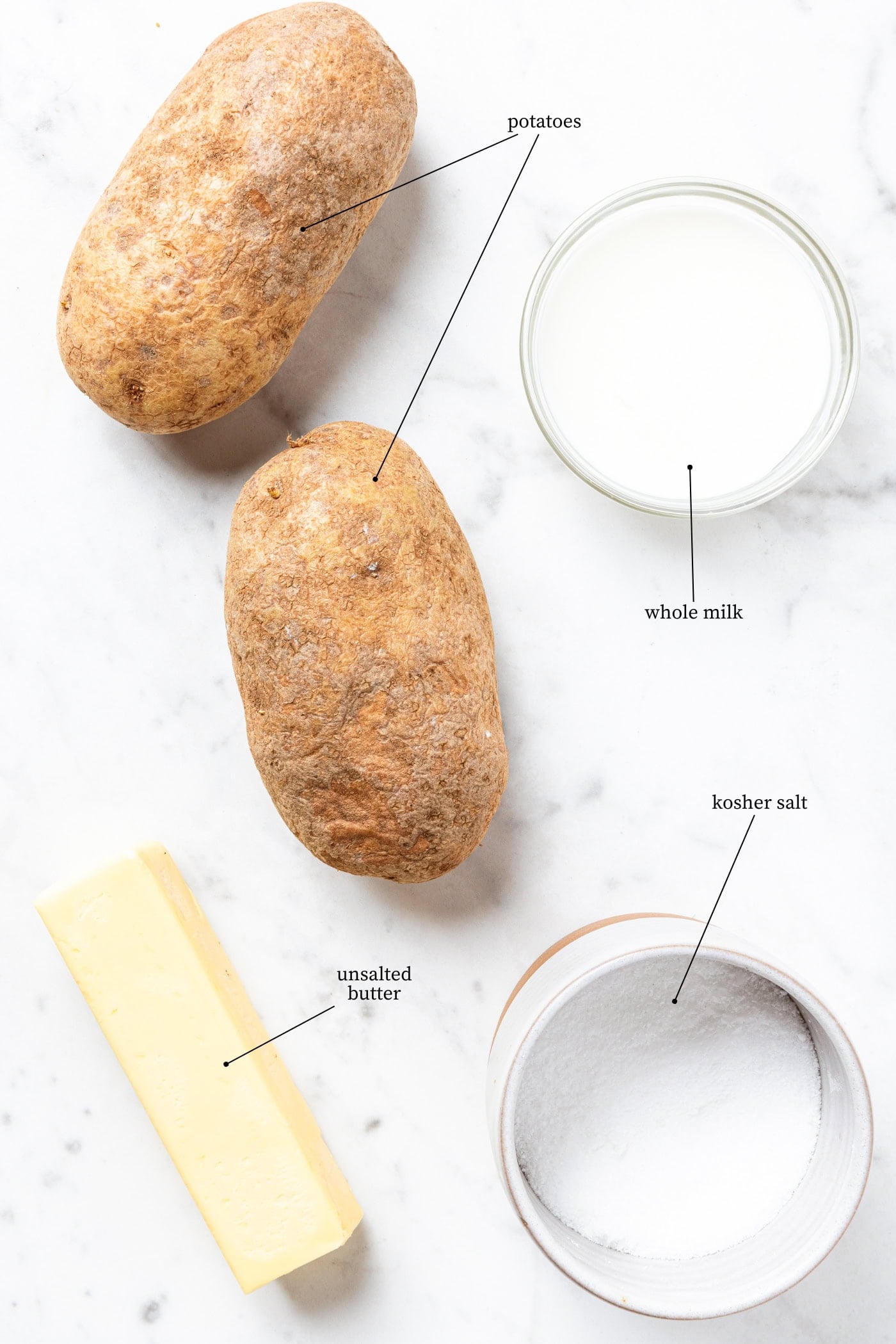 White marble surface filled with ingredients for mashed potatoes including potatoes, salt, butter, and milk.
