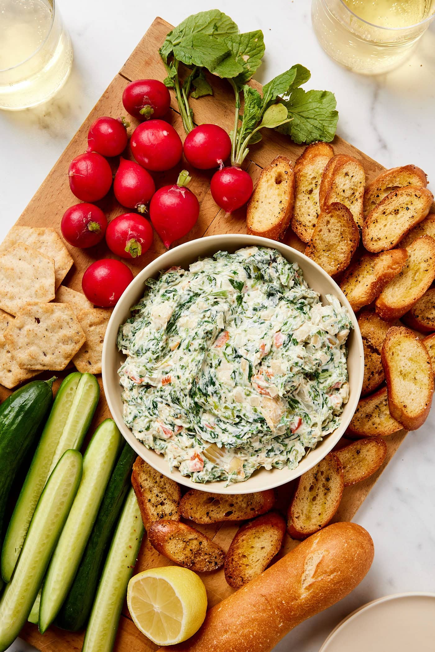 White bowl filled with spinach artichoke dip with fresh vegetables and crostini around on a wooden cutting board.