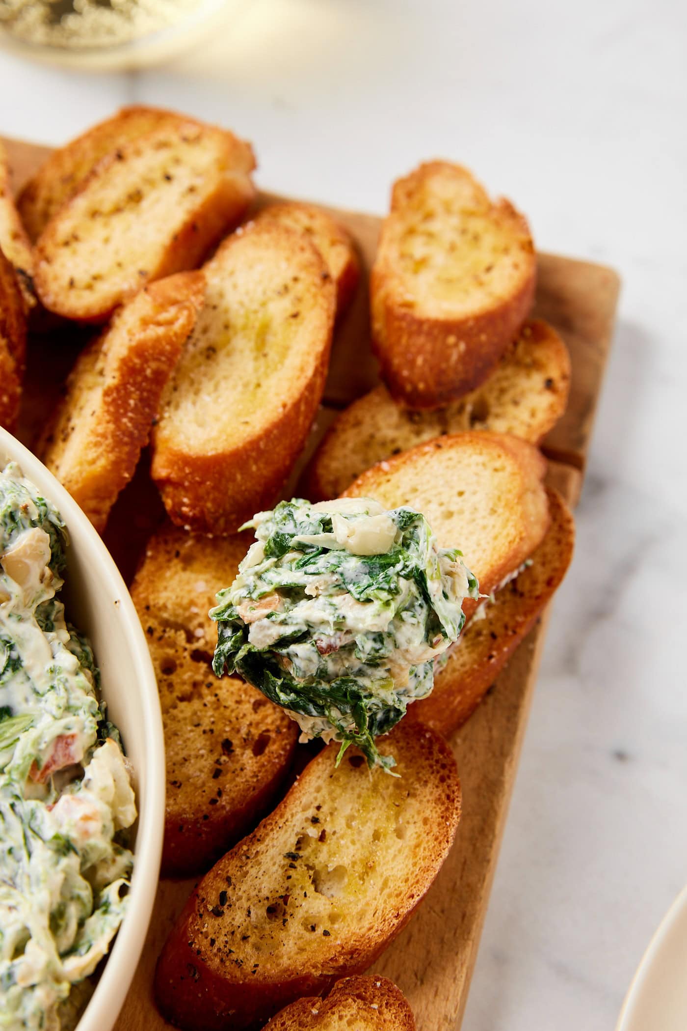 Crostini sitting on wooden cutting board with scoop of artichoke dip on one piece.