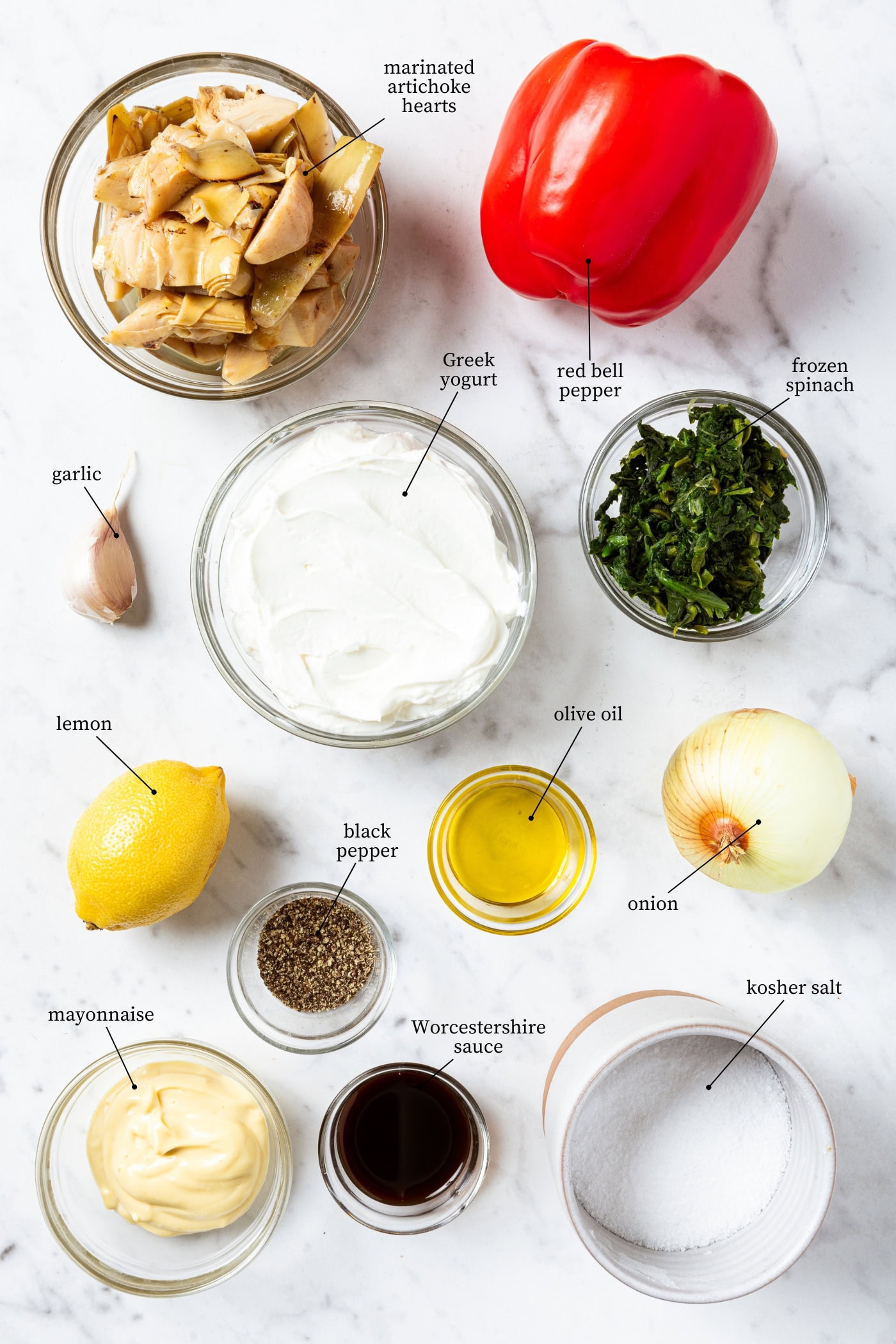 White marble surface filled with ingredients needed for spinach artichoke dip including artichokes, Greek yogurt, bell pepper, spinach, and more.