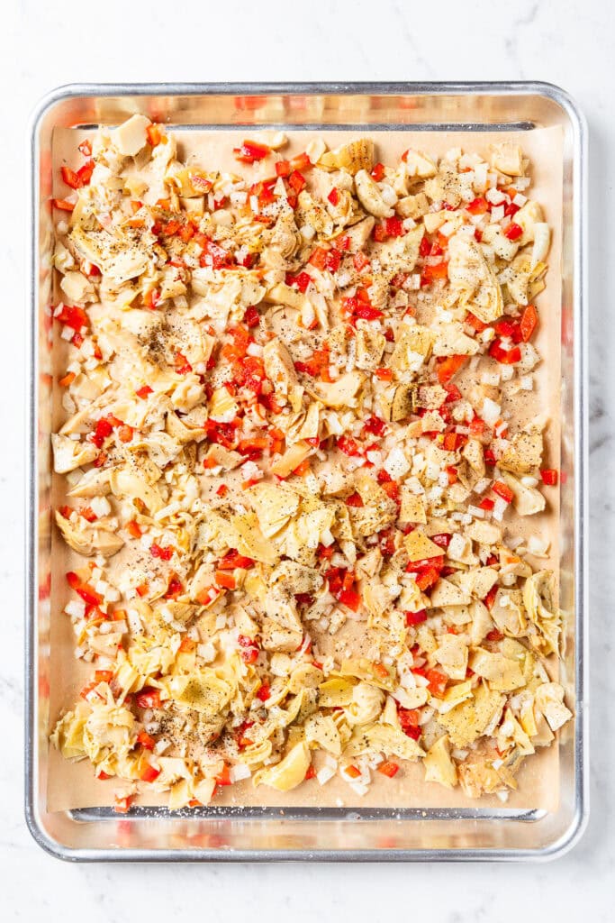 Artichoke hearts, onions, and red peppers on a pan with olive oil before roasting.