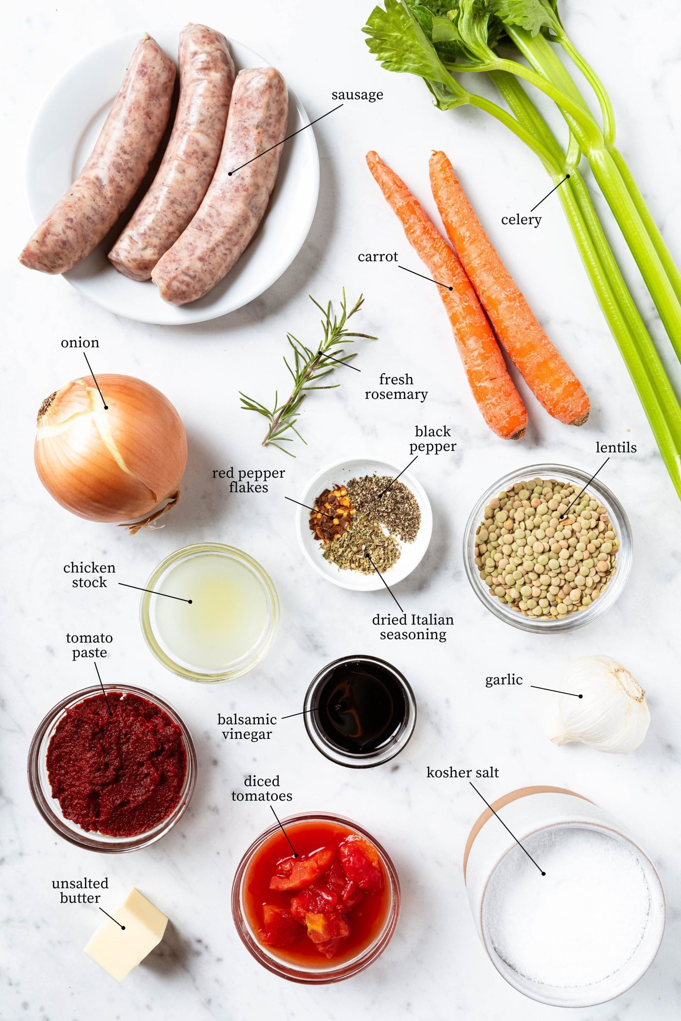 White marble surface filled with ingredients for sausage lentil soup including vegetables, spices, and more.