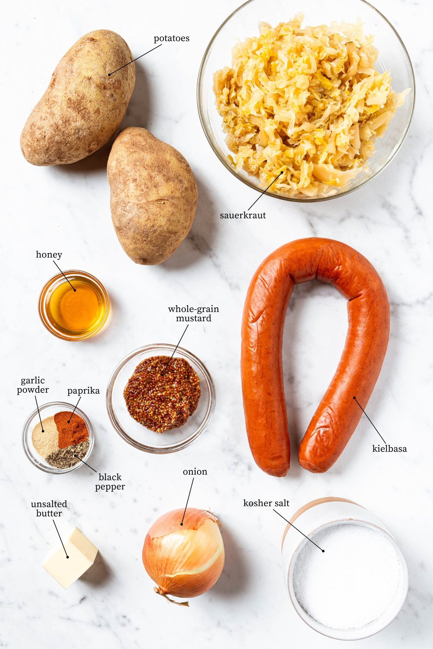 White marble surface filled with ingredients for kielbasa and sauerkraut including potatoes, spices, honey, and more.