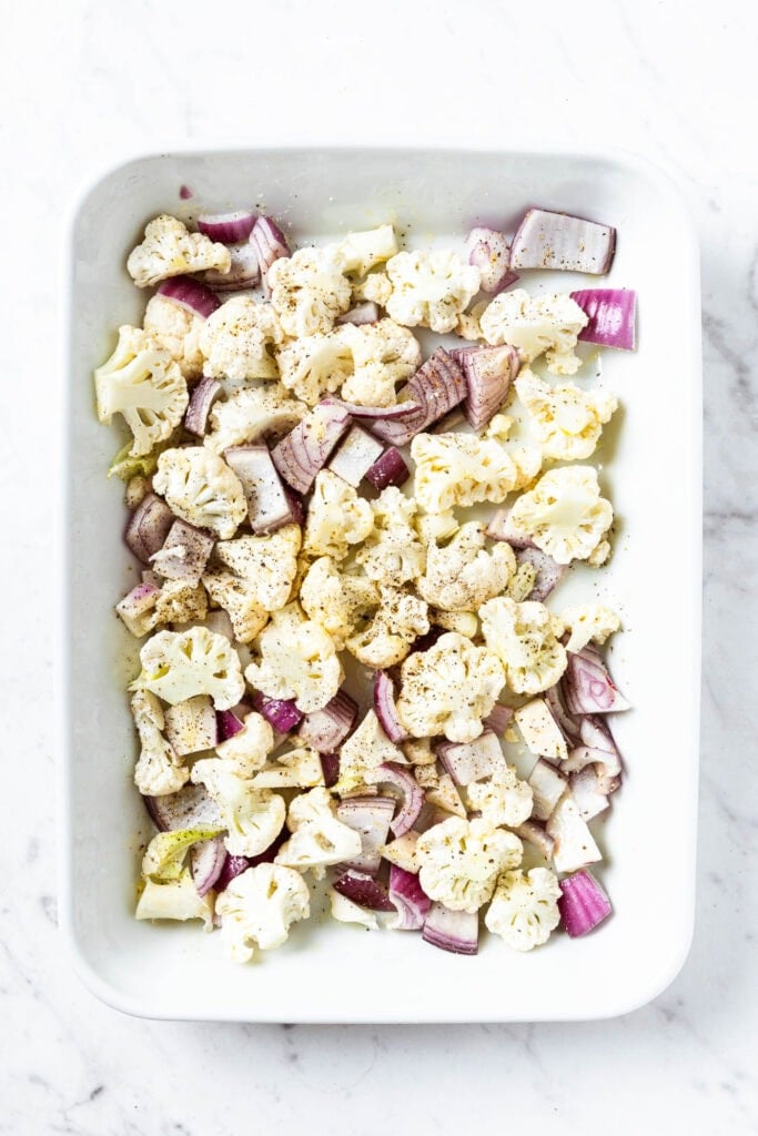 Cauliflower florets and red onion pieces scattered in a baking dish prior to roasting.