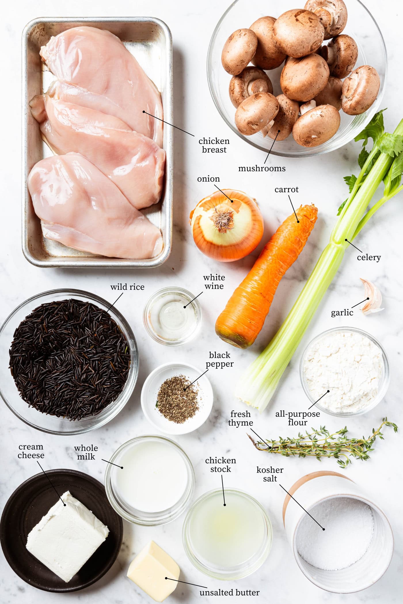 White marble surface filled with ingredients for chicken wild rice soup including raw chicken breast, vegetables, rice, spices, and more.