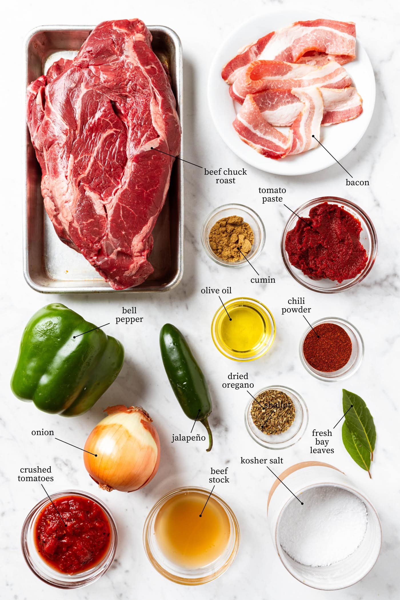White marble surface filled with ingredients for chili con carne recipe including vegetables, spices, bacon, beef, and more.