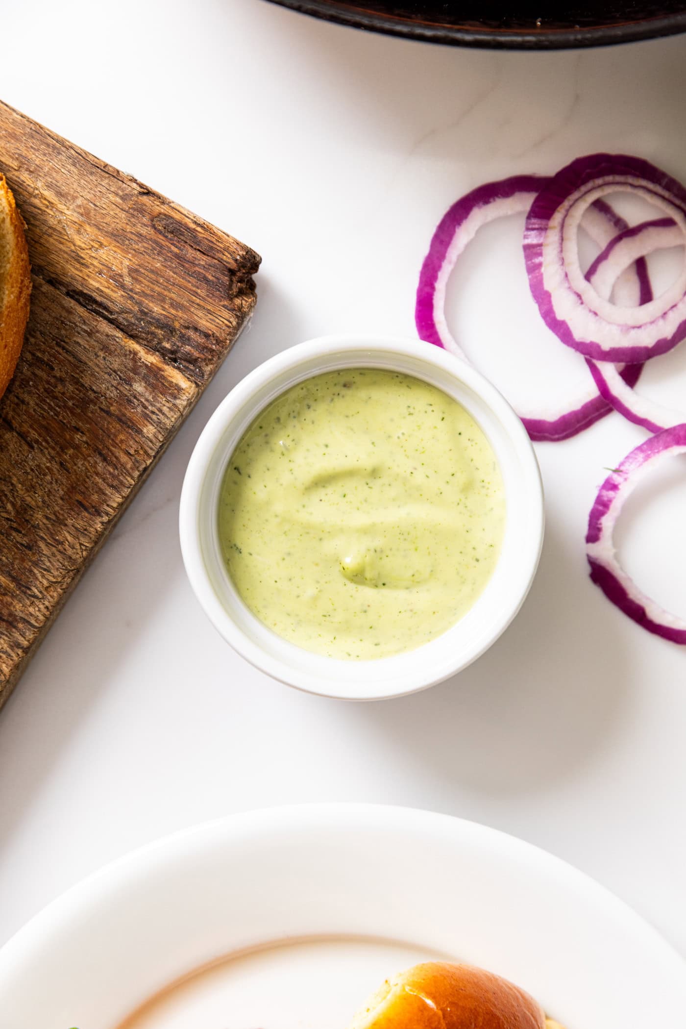 White bowl filled with dill aioli with wooden cutting board and red onions.