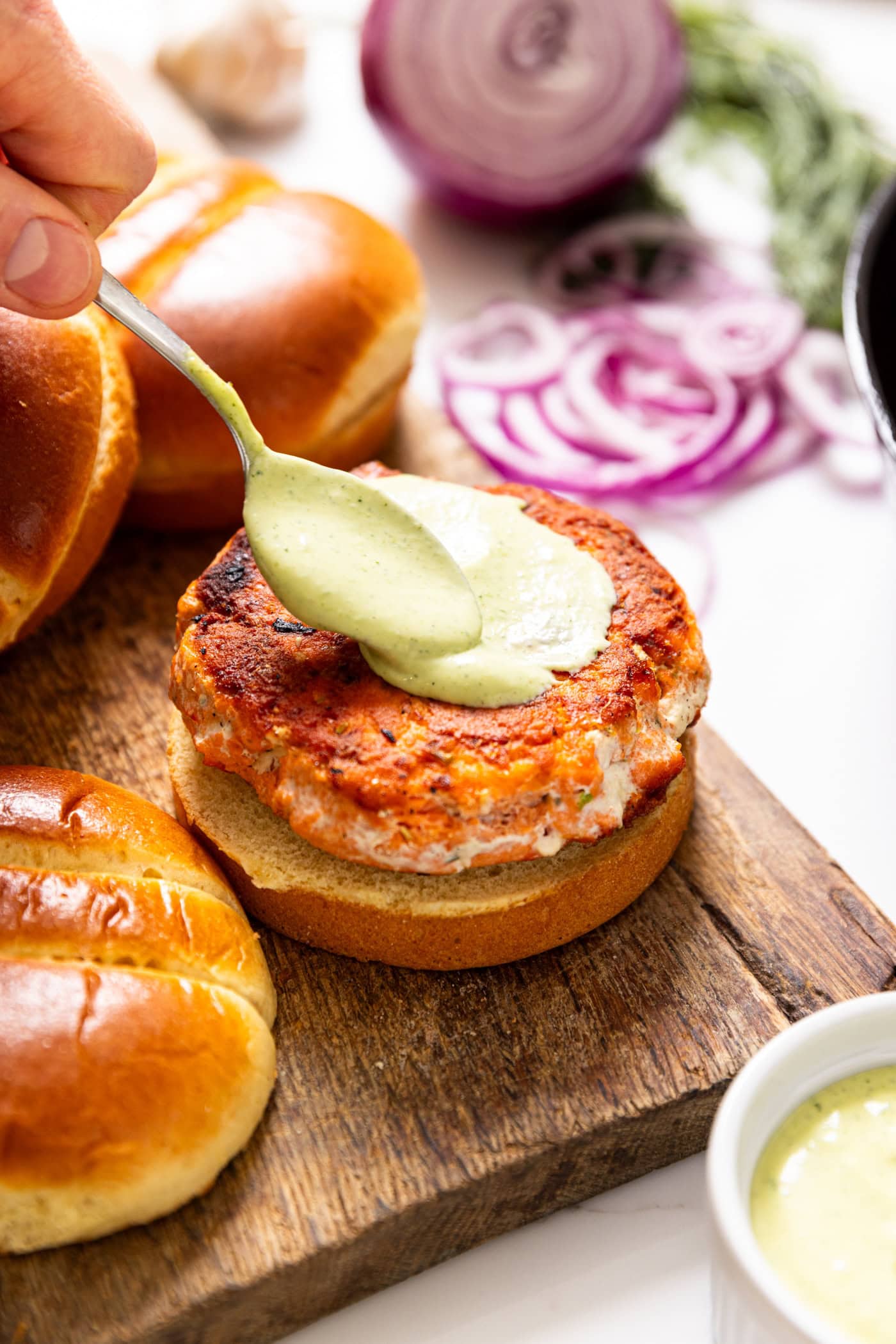 Spoon putting dill aioli sauce on top of salmon burger sitting on a golden bun.