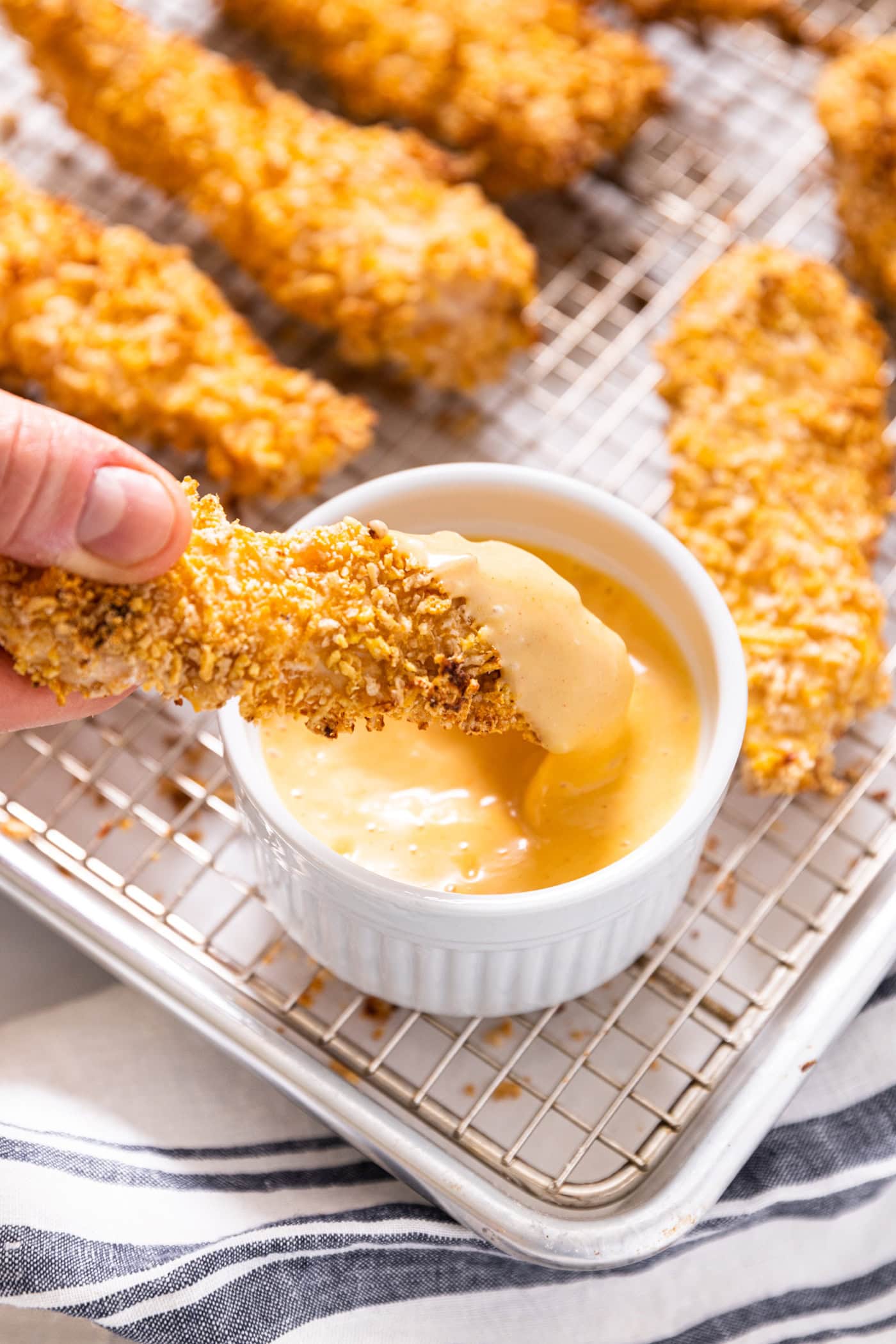 Hand holding chicken tender dipping into honey mustard sauce in a white bowl.