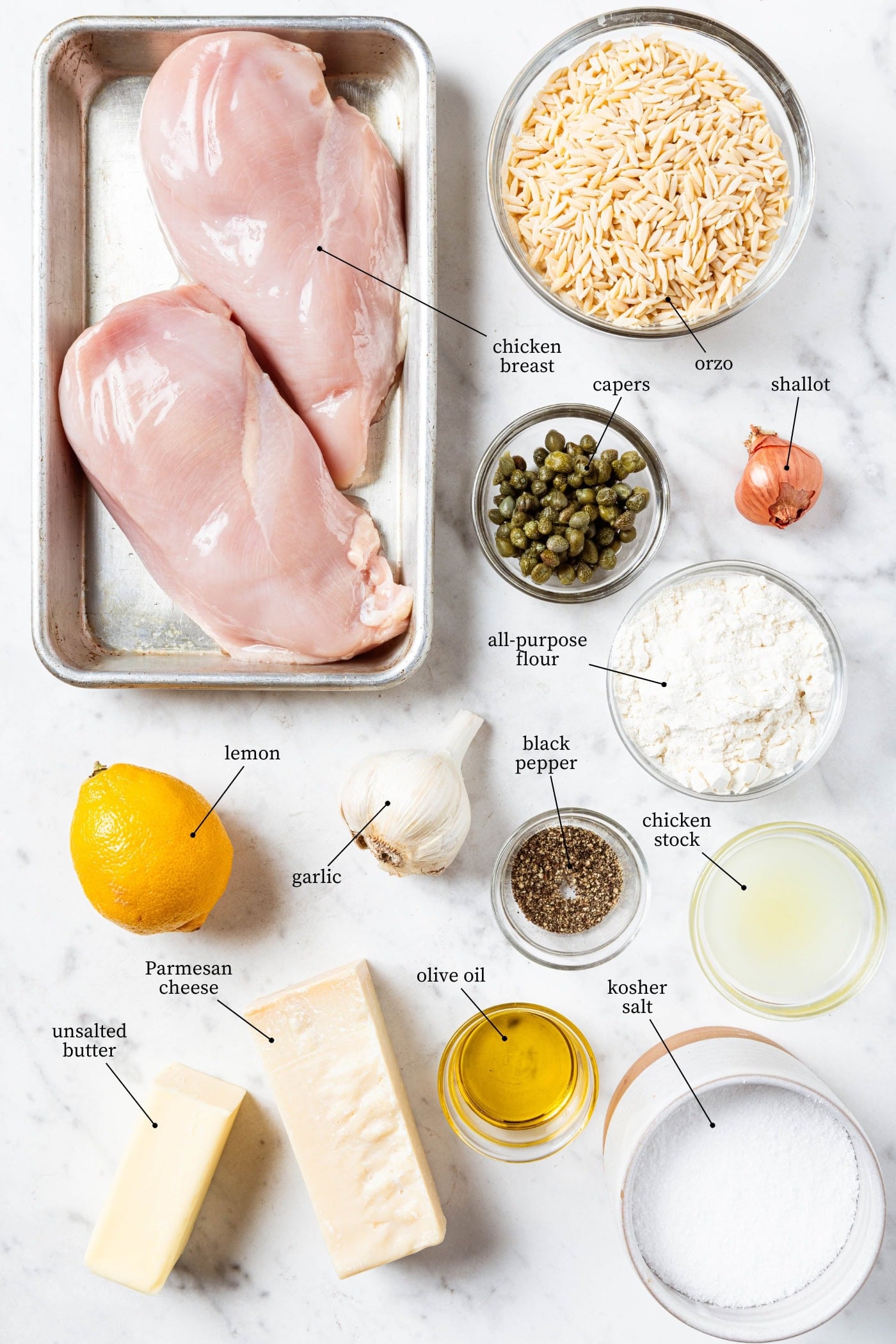 White marble surface filled with ingredients for chicken piccata including chicken breasts, capers, lemon, orzo, and more.