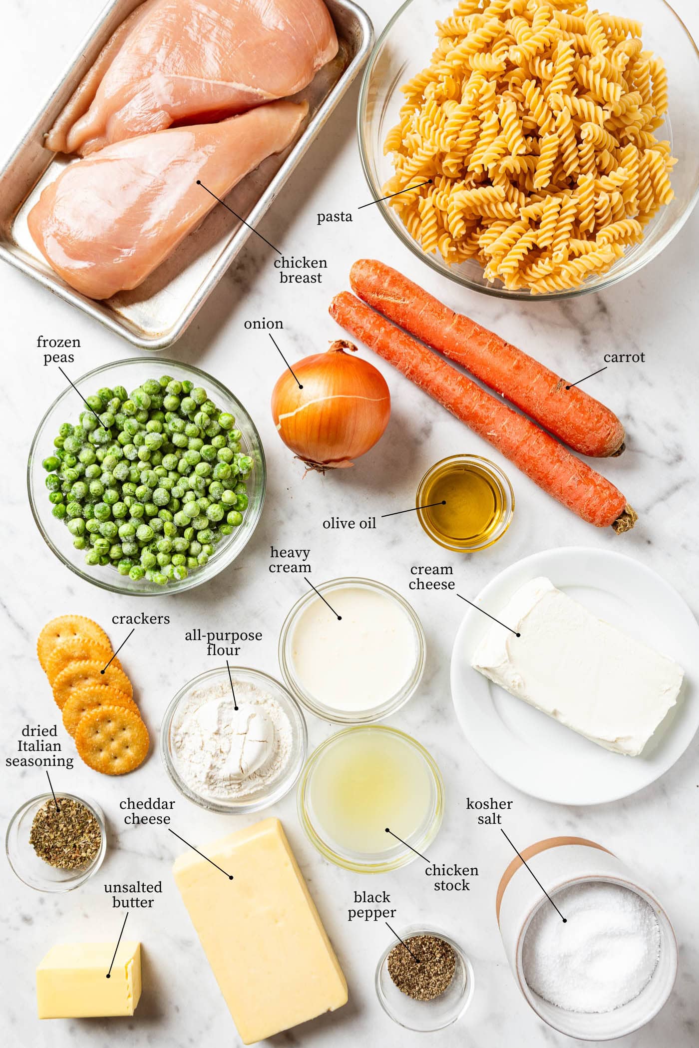 White marble surface filled with ingredients for chicken pot pie casserole including chicken breasts, carrots, onion, peas, pasta, and more.