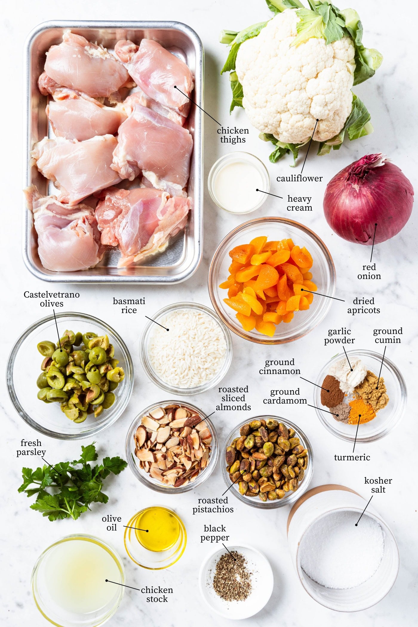 White marble surface filled with ingredients with rice pilaf including raw chicken, rice, nuts, olives, spices, herbs, and more.