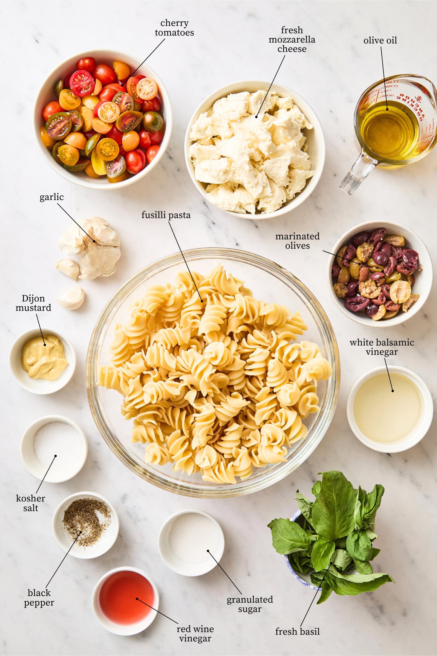 White marble surface filled with ingredients needed for caprese pasta salad including pasta, tomatoes, cheese, spices, and more.