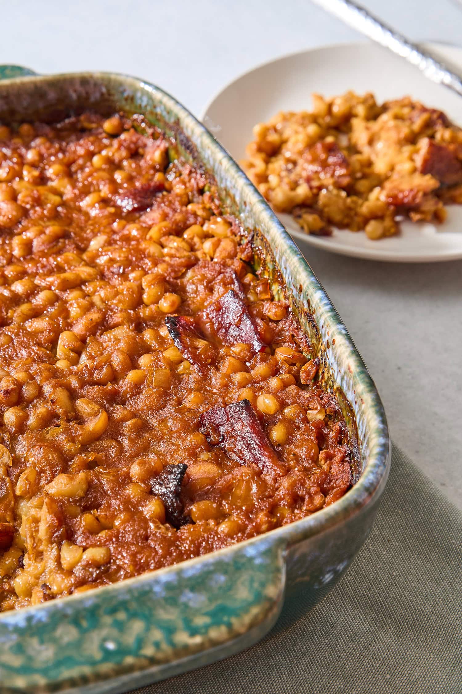 Baking dish with baked beans with bacon on top with plate filled with a serving.