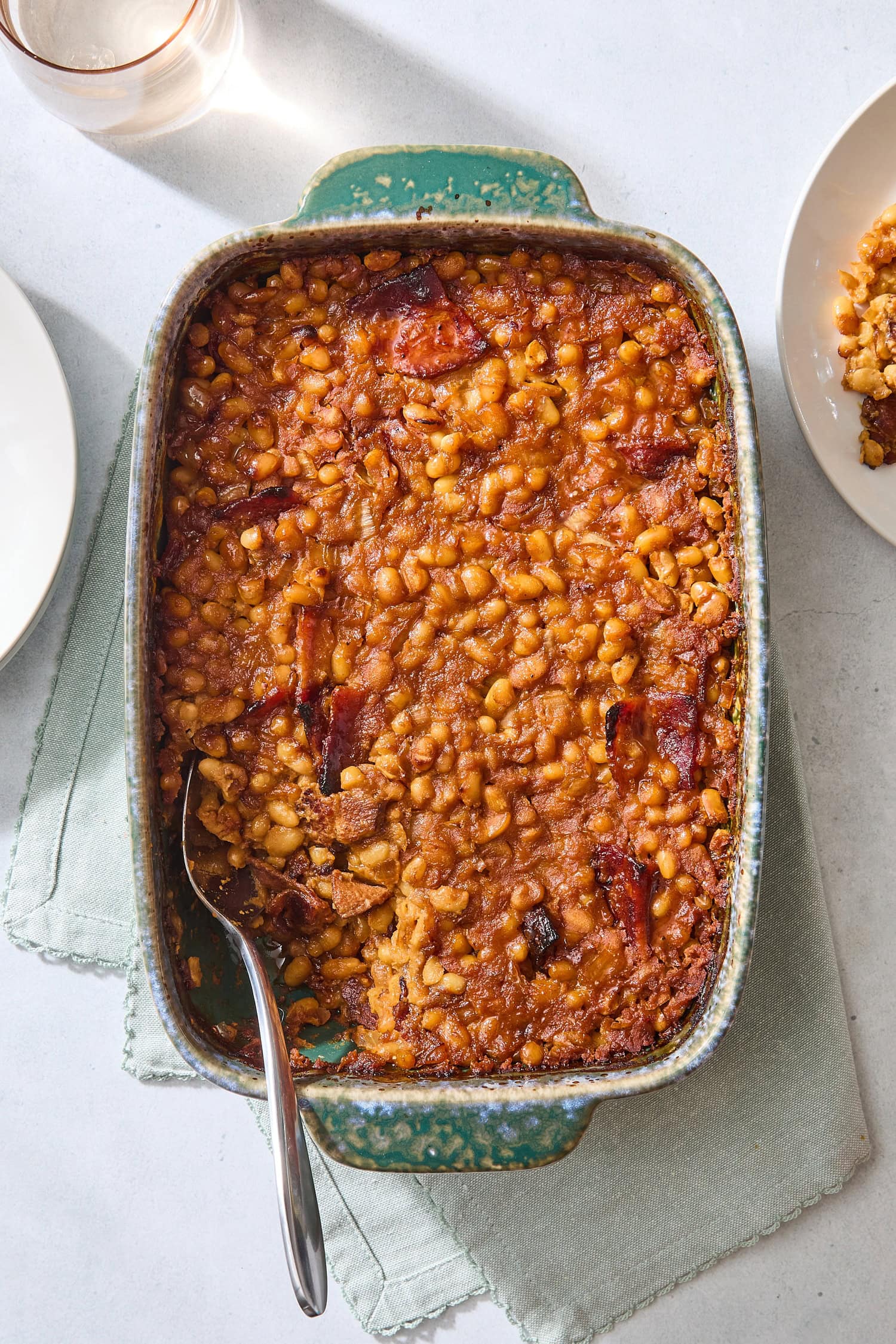 Pan filled with homemade baked beans with pieces of bacon on top with plates to the side.