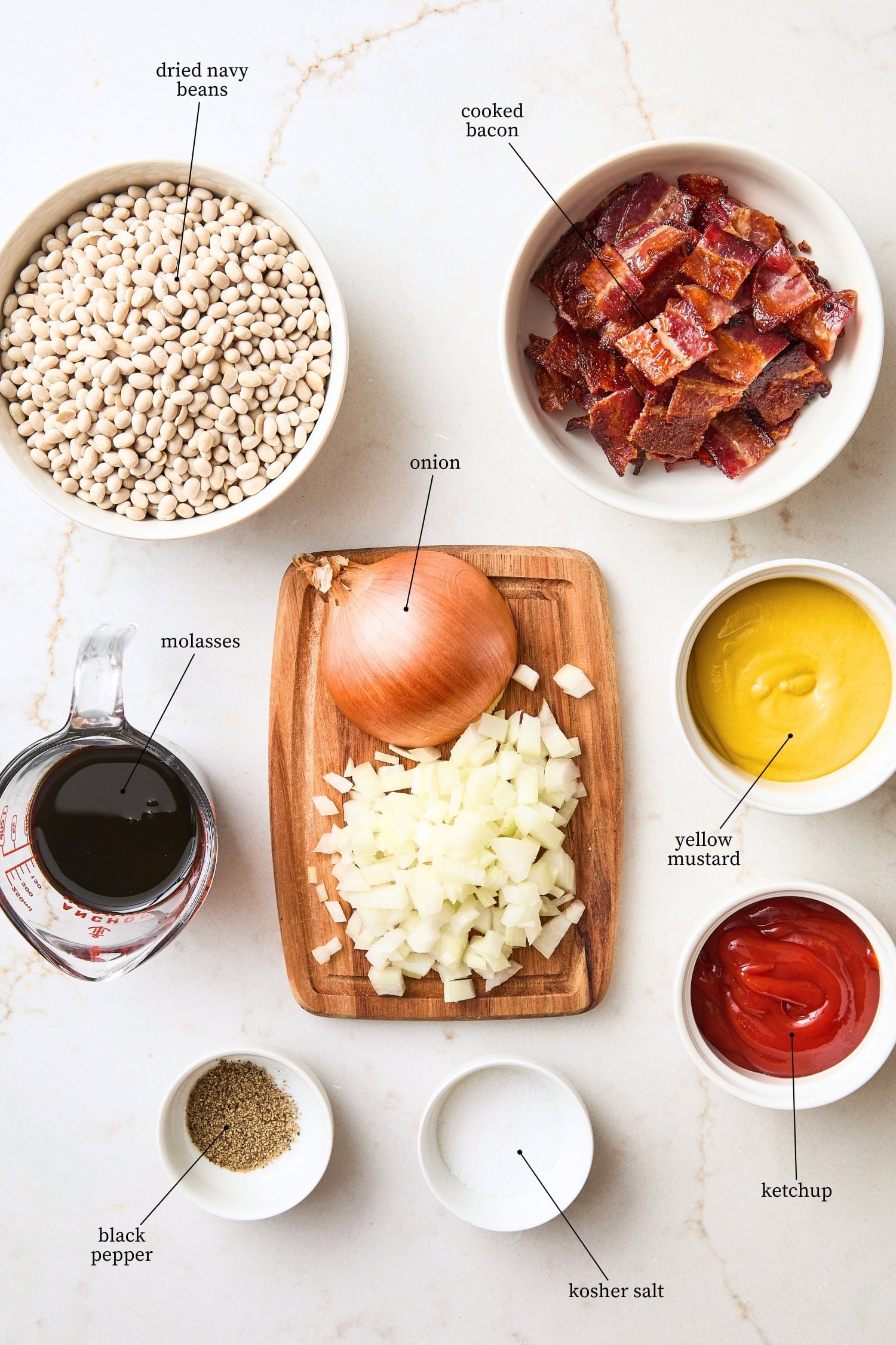 White surface filled with ingredients needed for homemade baked beans including beans, ketchup, mustard, bacon, and more.