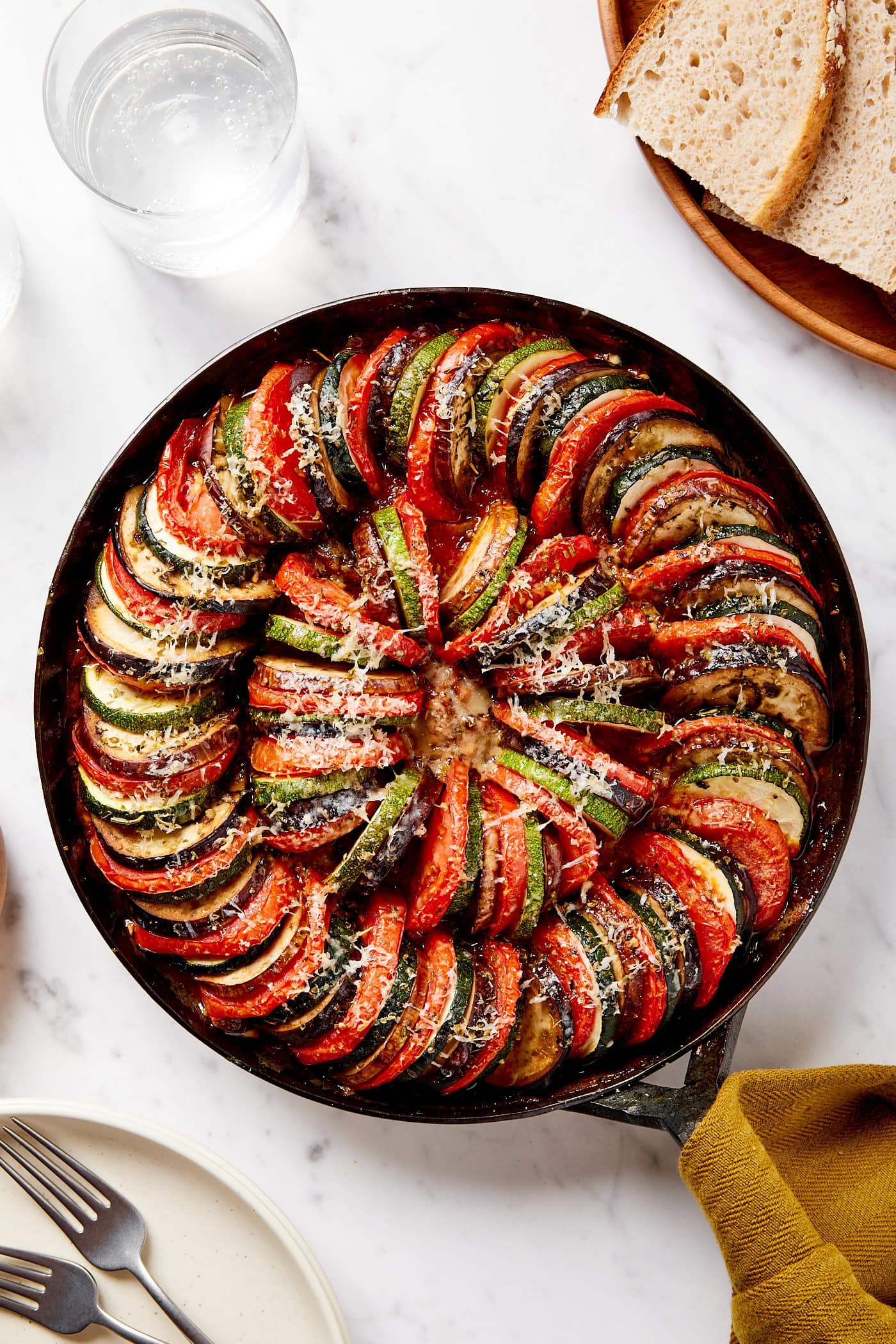 Black skillet with cooked ratatouille on white marble surface with bread to the side.