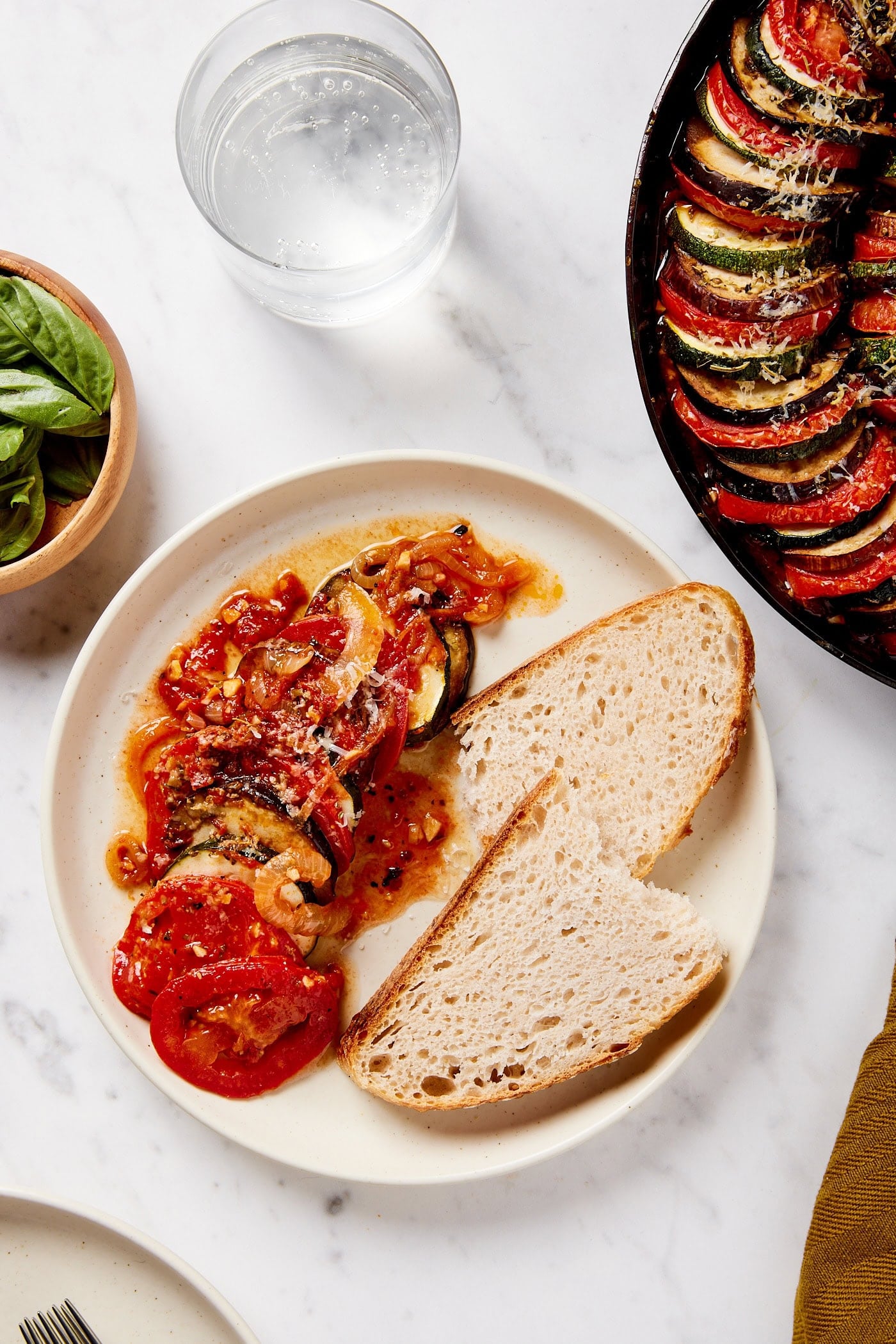 White plate with crusty bread and serving of ratatouille on white marble surface.