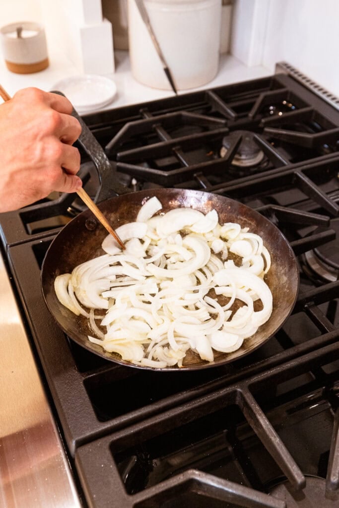 Skillet with wooden spoon stirring raw sliced onions.