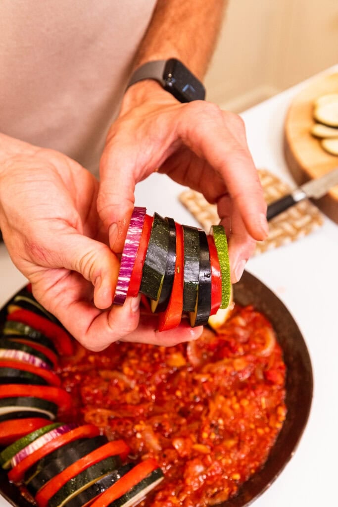 Slices of vegetables being placed in a skillet with sauce for ratatouille.