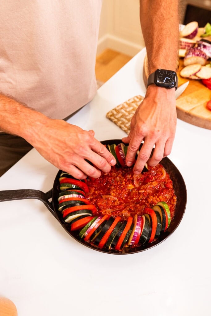 Slices of vegetables for ratatouille being arranged in a skillet with sauce.