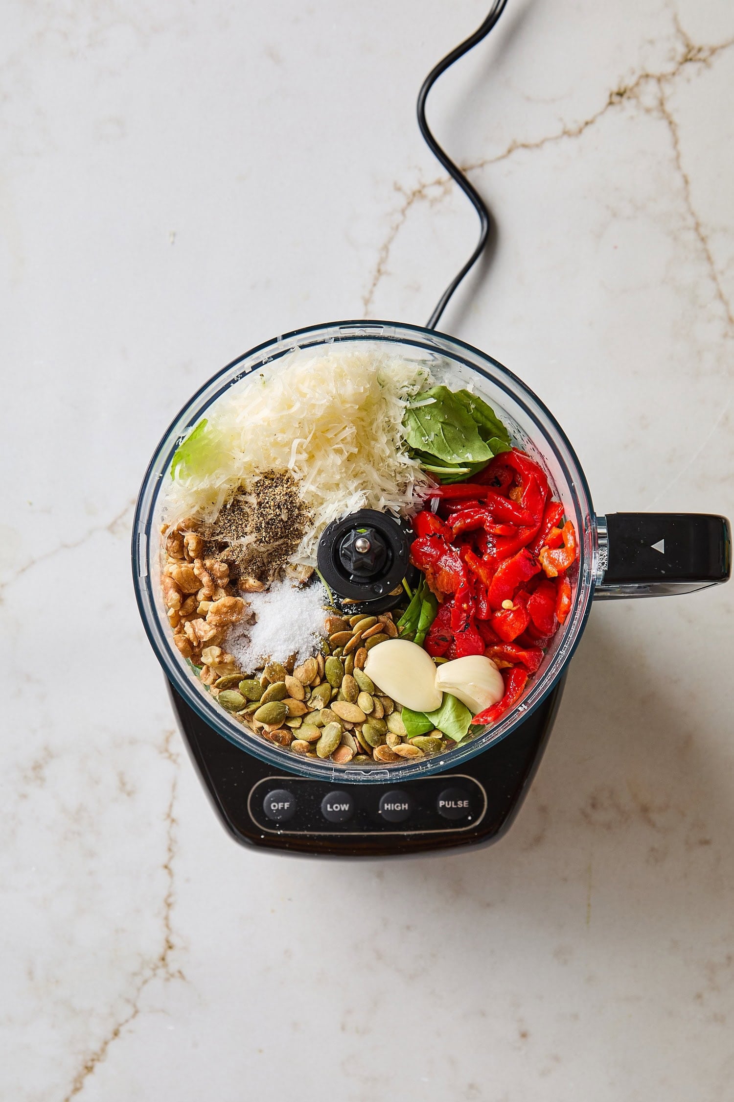 Food processor filled with whole ingredients for arugula pesto including arugula and roasted red peppers.