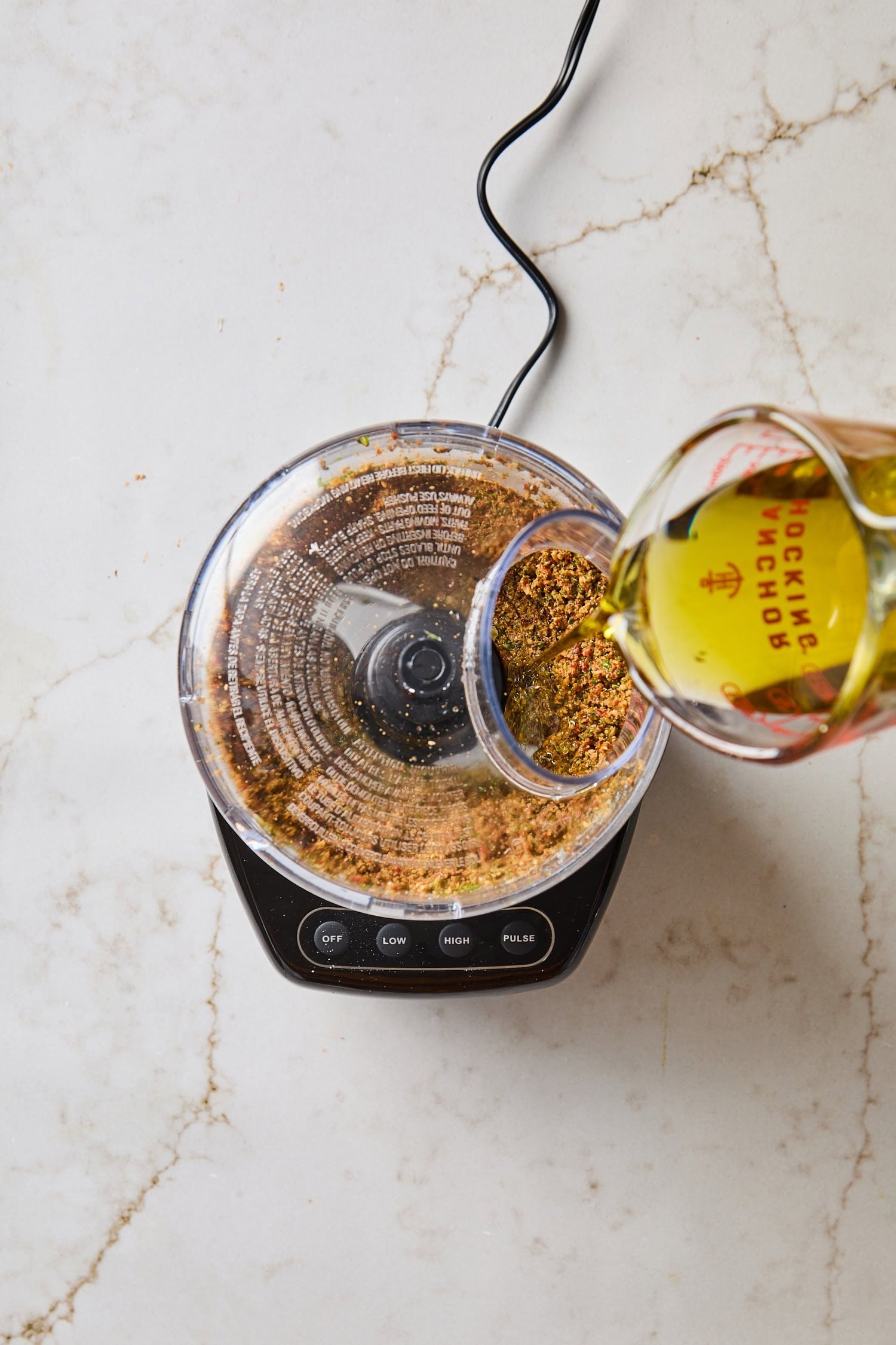 Measuring cup pouring olive oil into a food processor for pesto recipe.