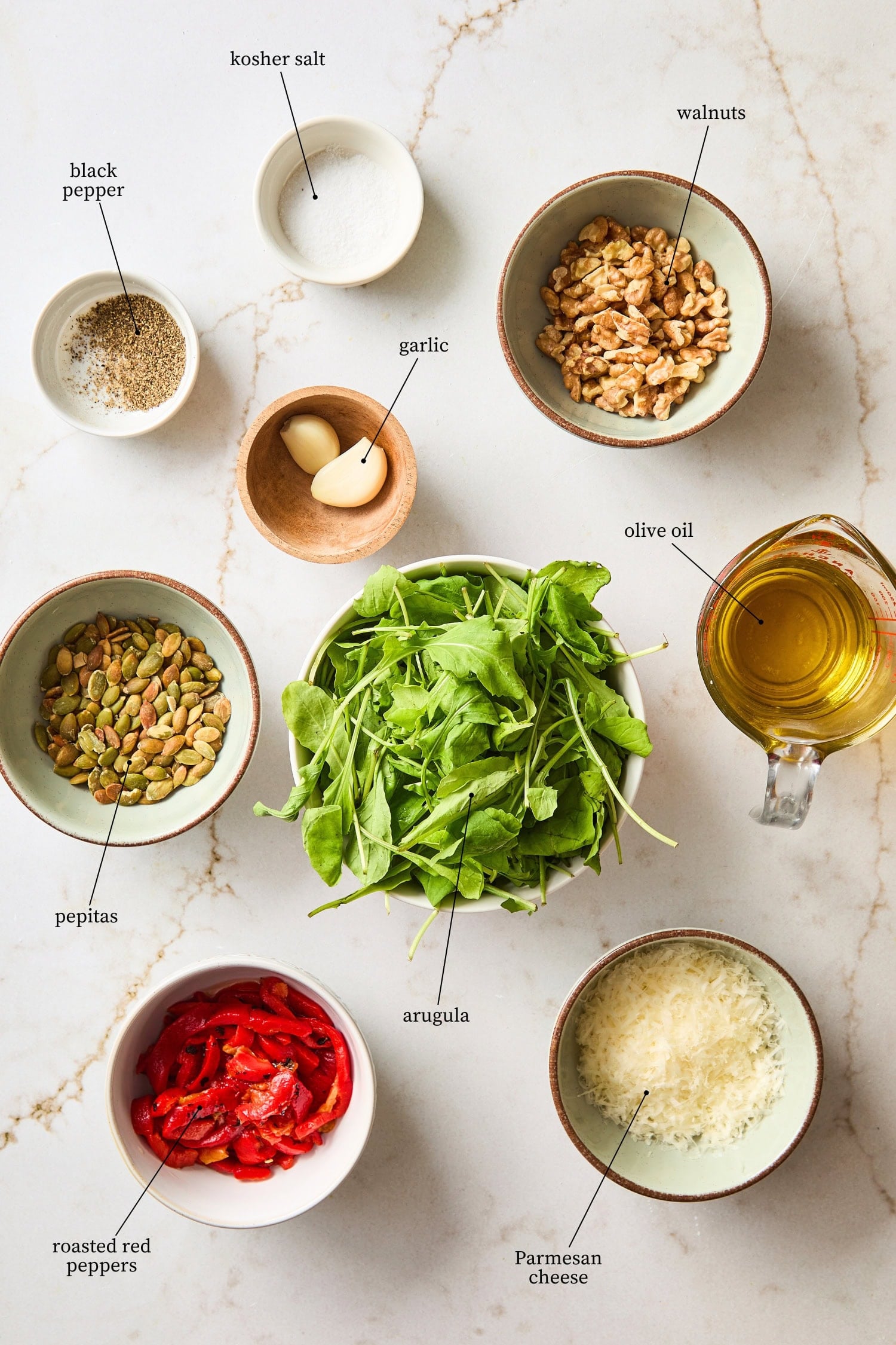 White countertop filled with ingredients for arugula pesto including arugula, walnuts, roasted red peppers, and more.