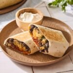 Tile surface with brown plate with chorizo breakfast burritos with sour cream.