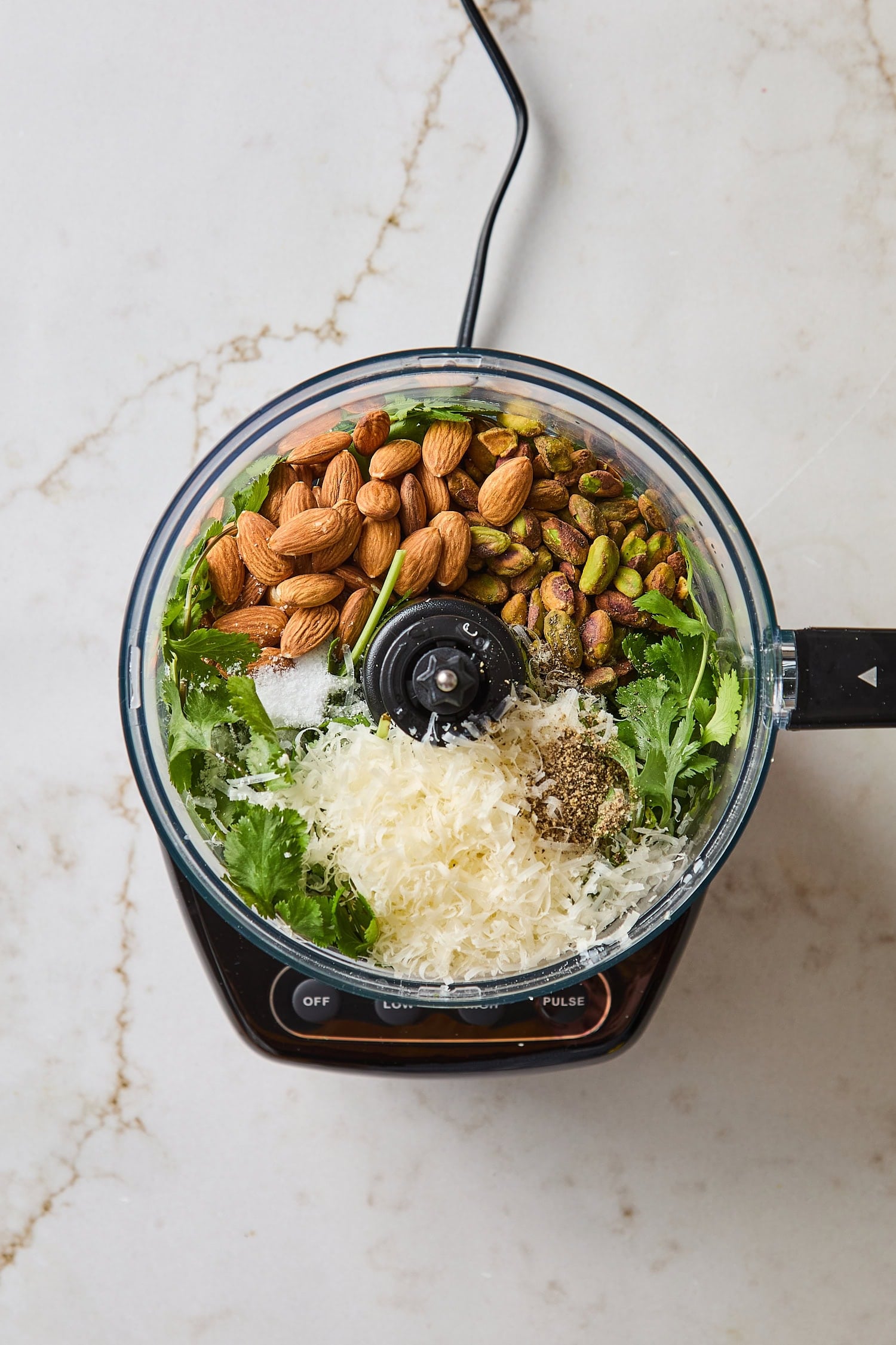 Food processor filled with ingredients for cilantro pesto including almonds, Parmesan cheese, and cilantro.