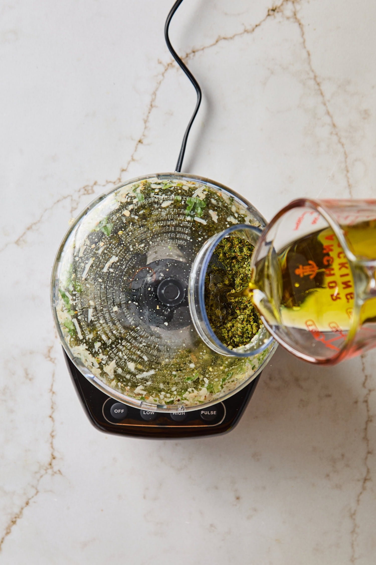 Measuring cup pouring olive oil into a food processor with green colored pesto.