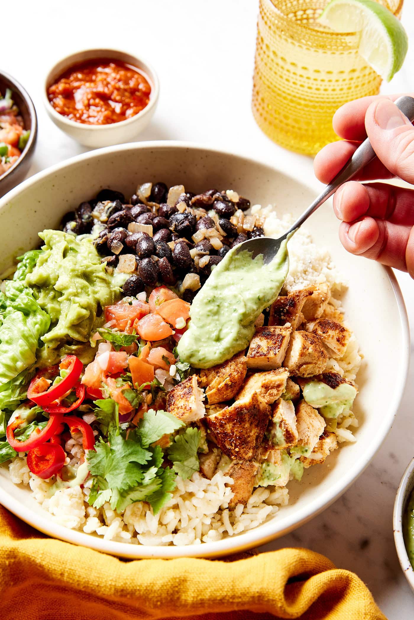 Hand spooning green dressing on top of chicken in bowl topped with rice and beans.